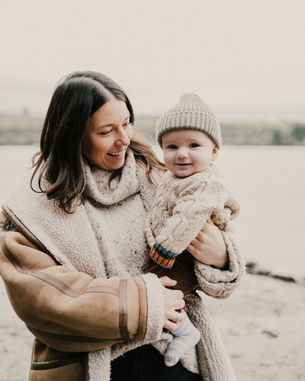 Mother smiling looking at baby in arms standing on Cornish beach.