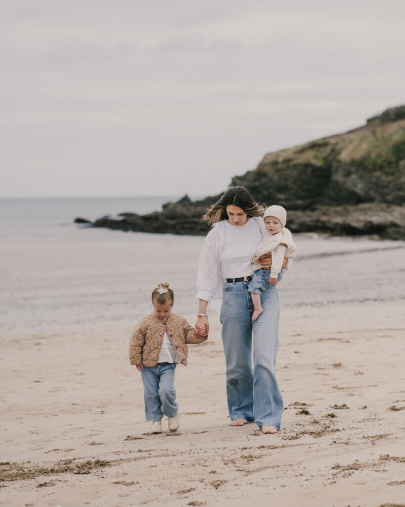 Mother walking hand in hand with her daughter while carrying her baby on Maenporth Beach