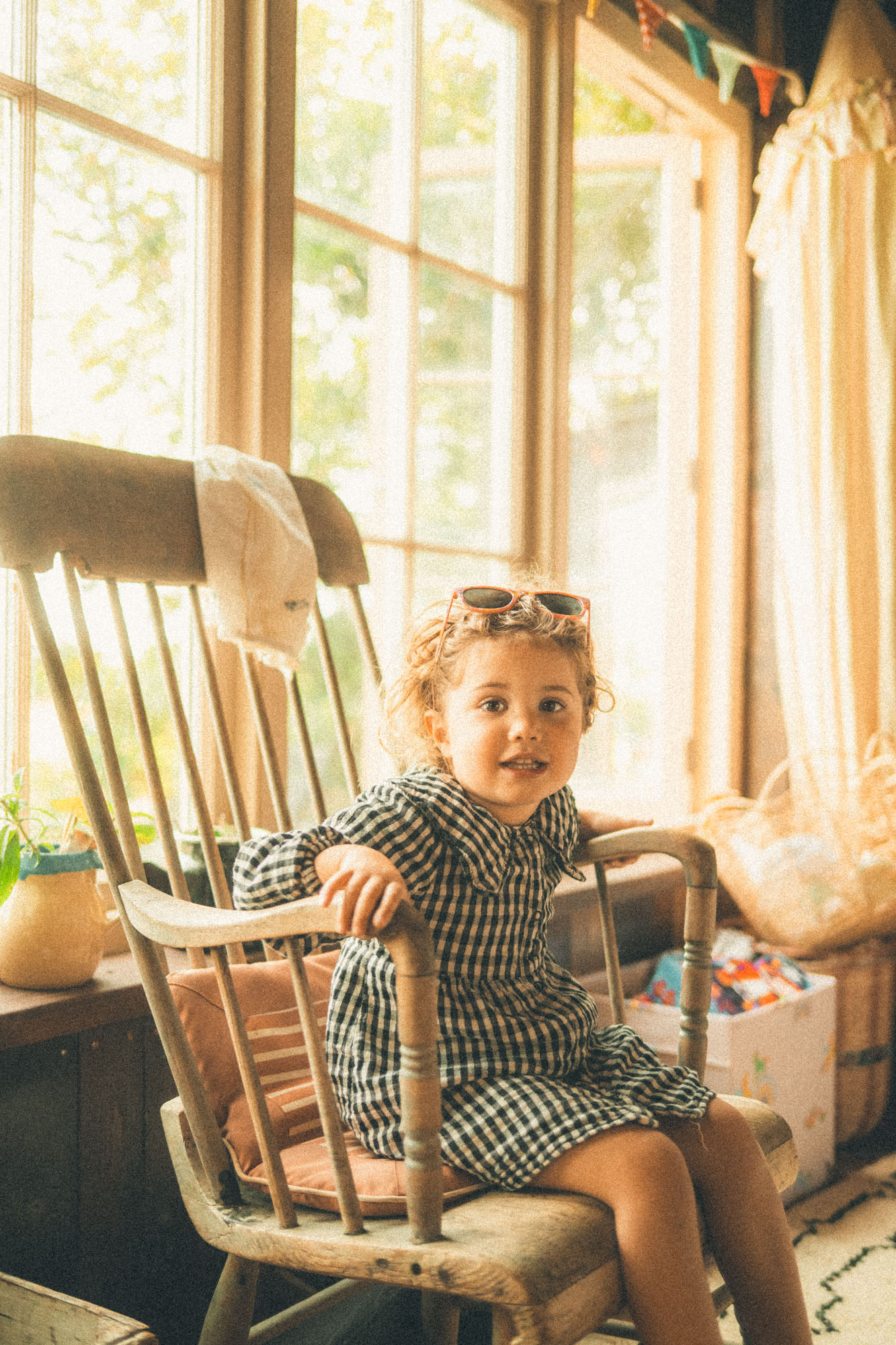 Young girl in a gingham dress sitting in a sunlit rocking chair at home