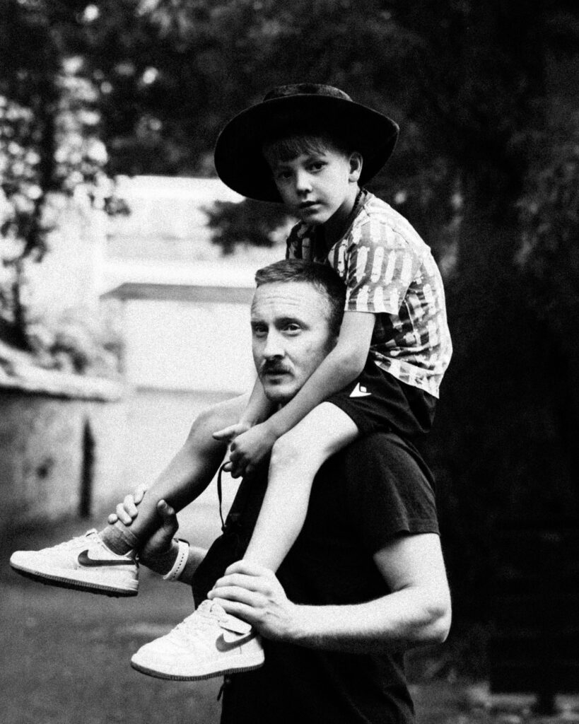 Black-and-white portrait of a boy wearing a hat, sitting on a man’s shoulders during a family photoshoot in Falmouth.