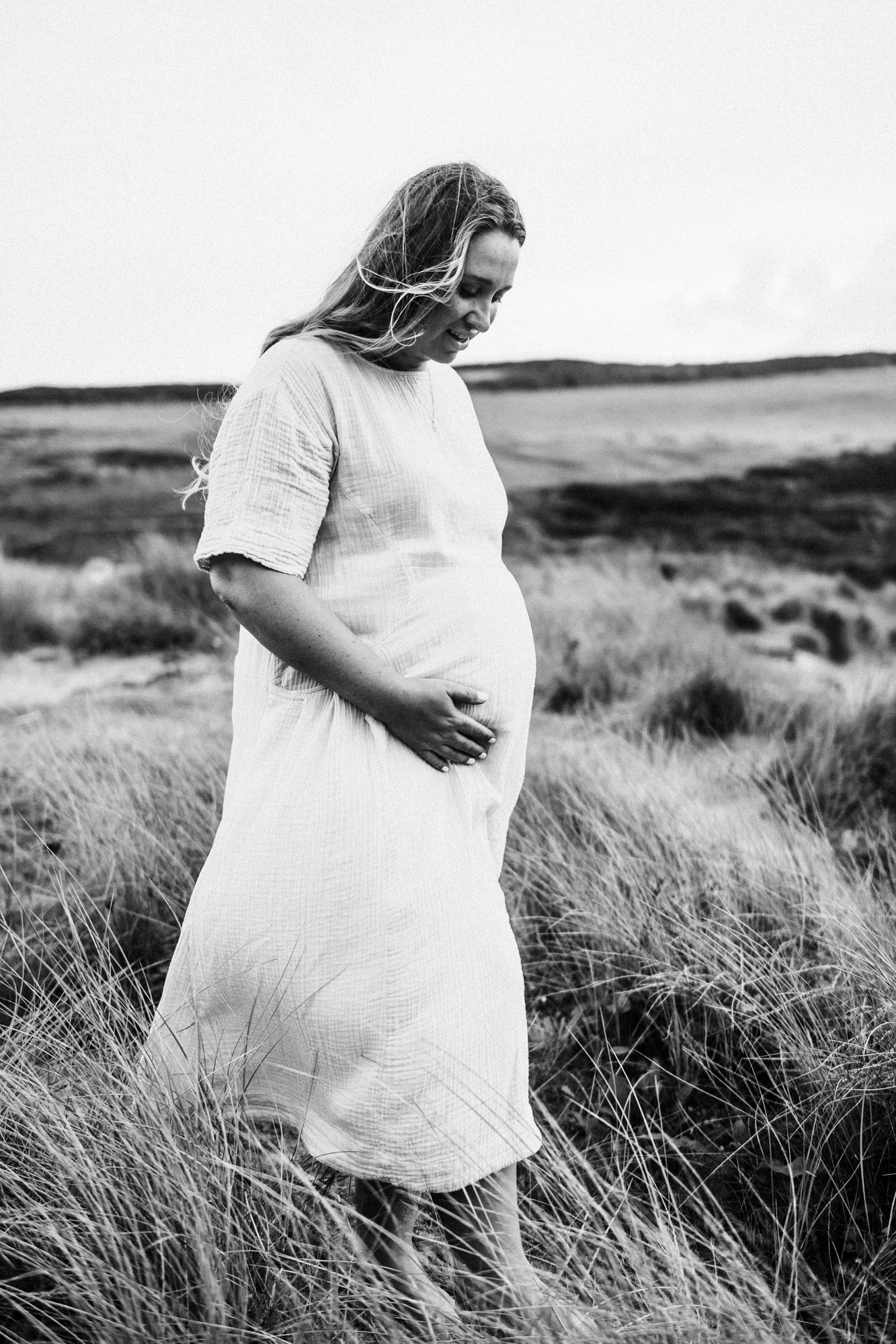 Black-and-white portrait of an expectant mother standing in long grass with a hand on her bump