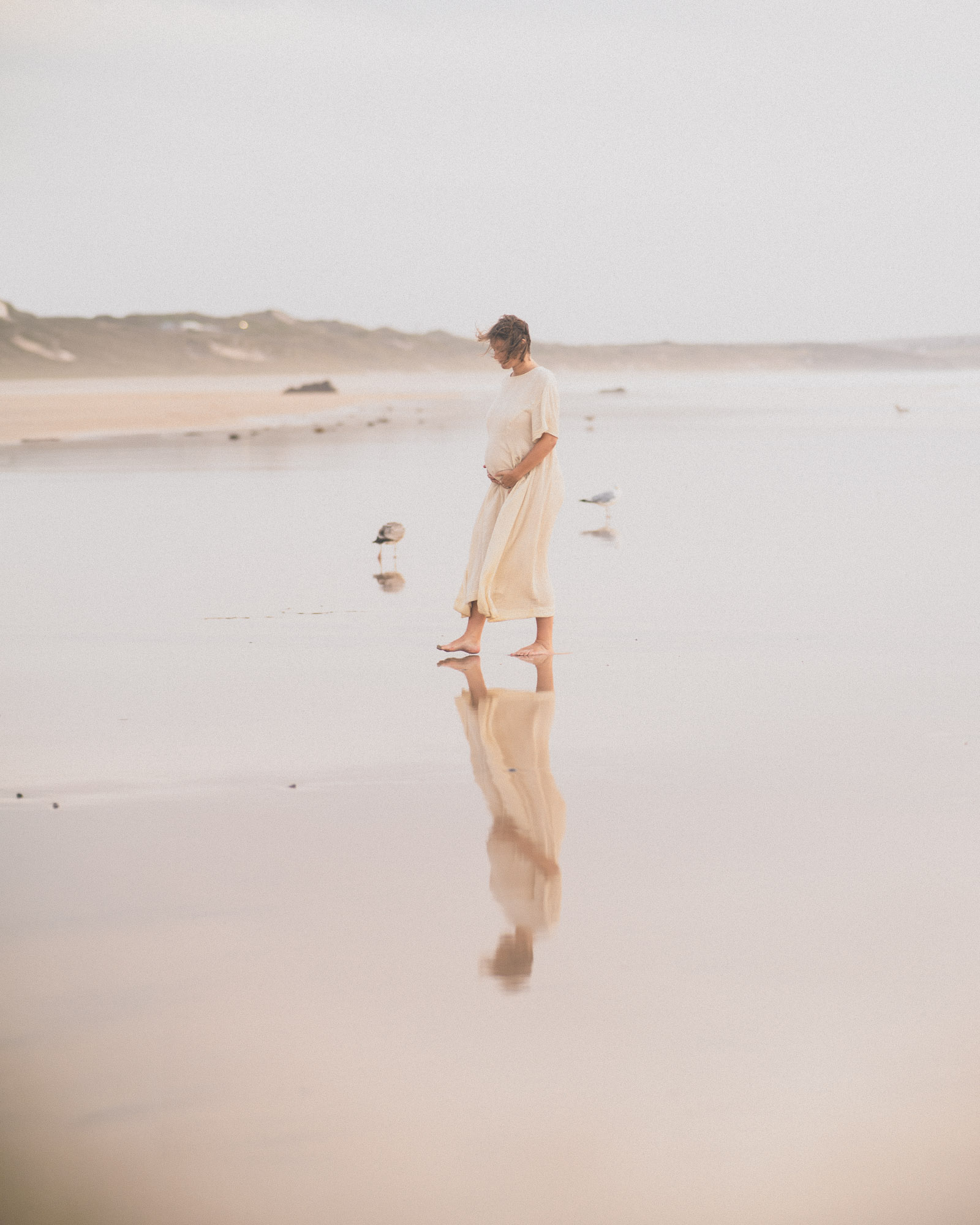 Expectant mother in a cream dress standing barefoot on wet sand with her reflection below